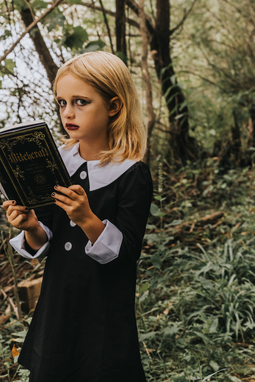 Girl in a black cotton dress with a white Peter Pan collar and cuffed sleeves, holding a witchcraft book in a forest.