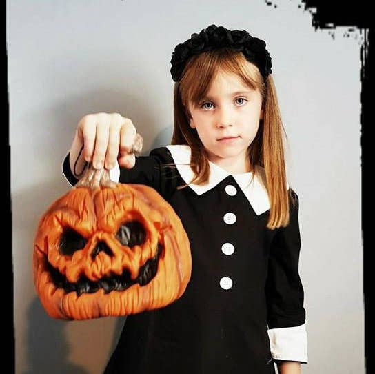 Girl in a black gothic dress with a white Peter Pan collar and cuffs, holding a carved pumpkin for Halloween.