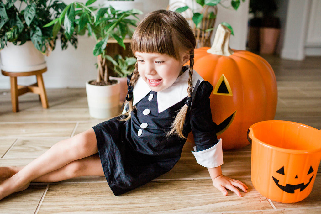 Girl in a black gothic cotton dress with a white Peter Pan collar and cuffed sleeves, sitting next to a Halloween pumpkin.